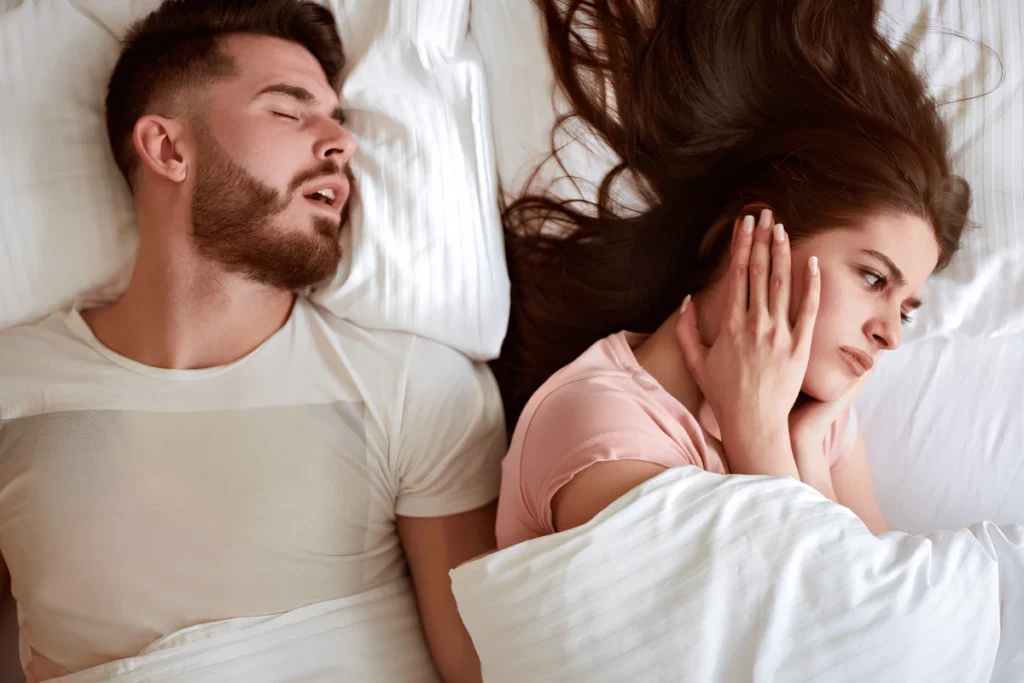 A man sleeping with his mouth open while snoring loudly, and his frustrated partner covering her ears beside him in bed.